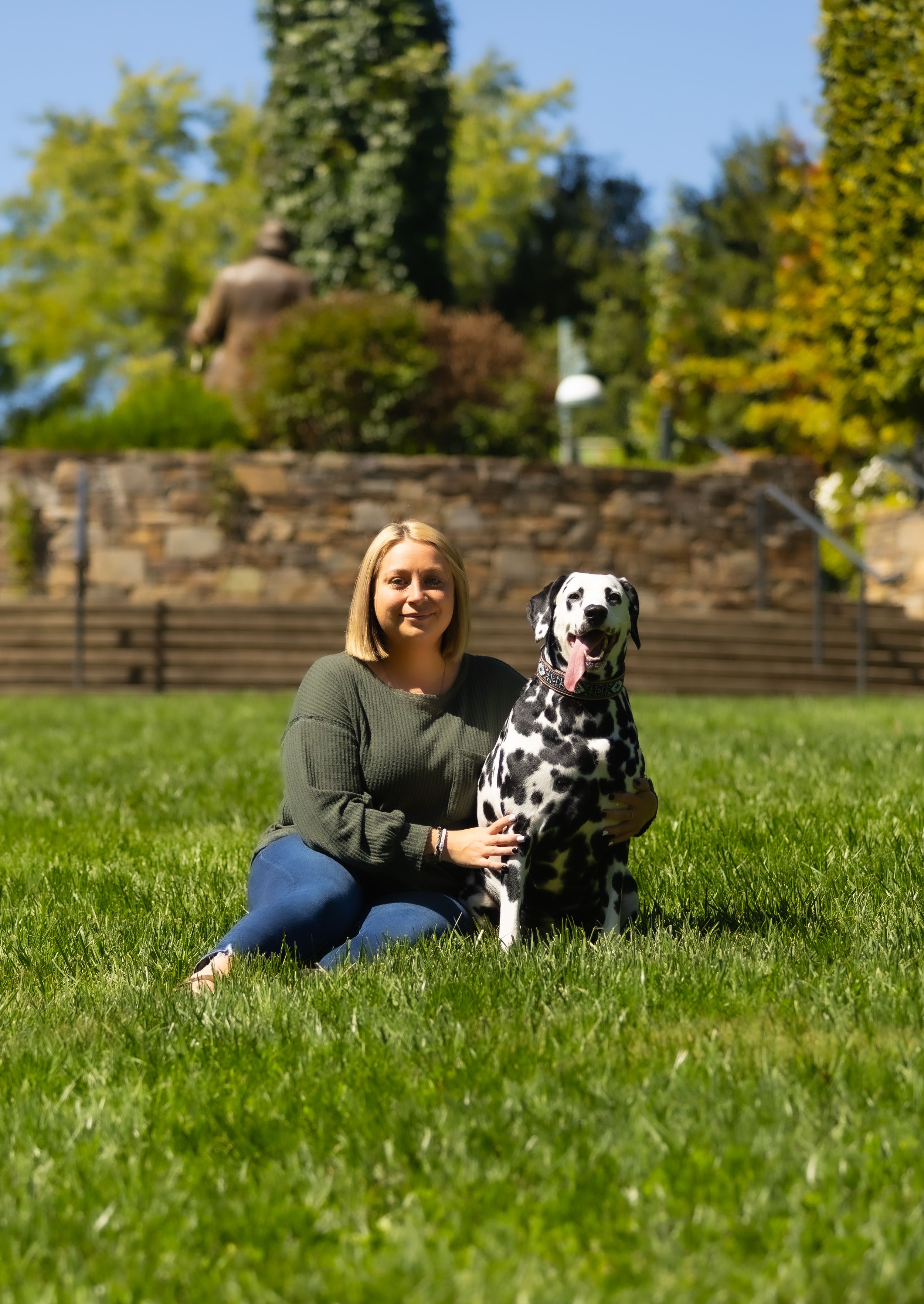 A Dalmatian dog sits on green grass with its companion on a sunny autumn day near stone steps.