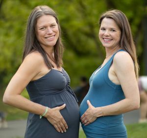 Two pregnant women wearing form-fitting tank tops in blue and gray stand together outdoors smiling in the sunlight.