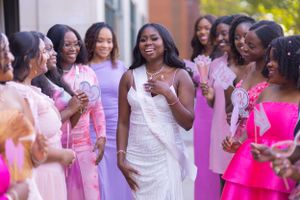 A group of wedding guests in pink and white dresses share joyful laughter at an outdoor celebration.