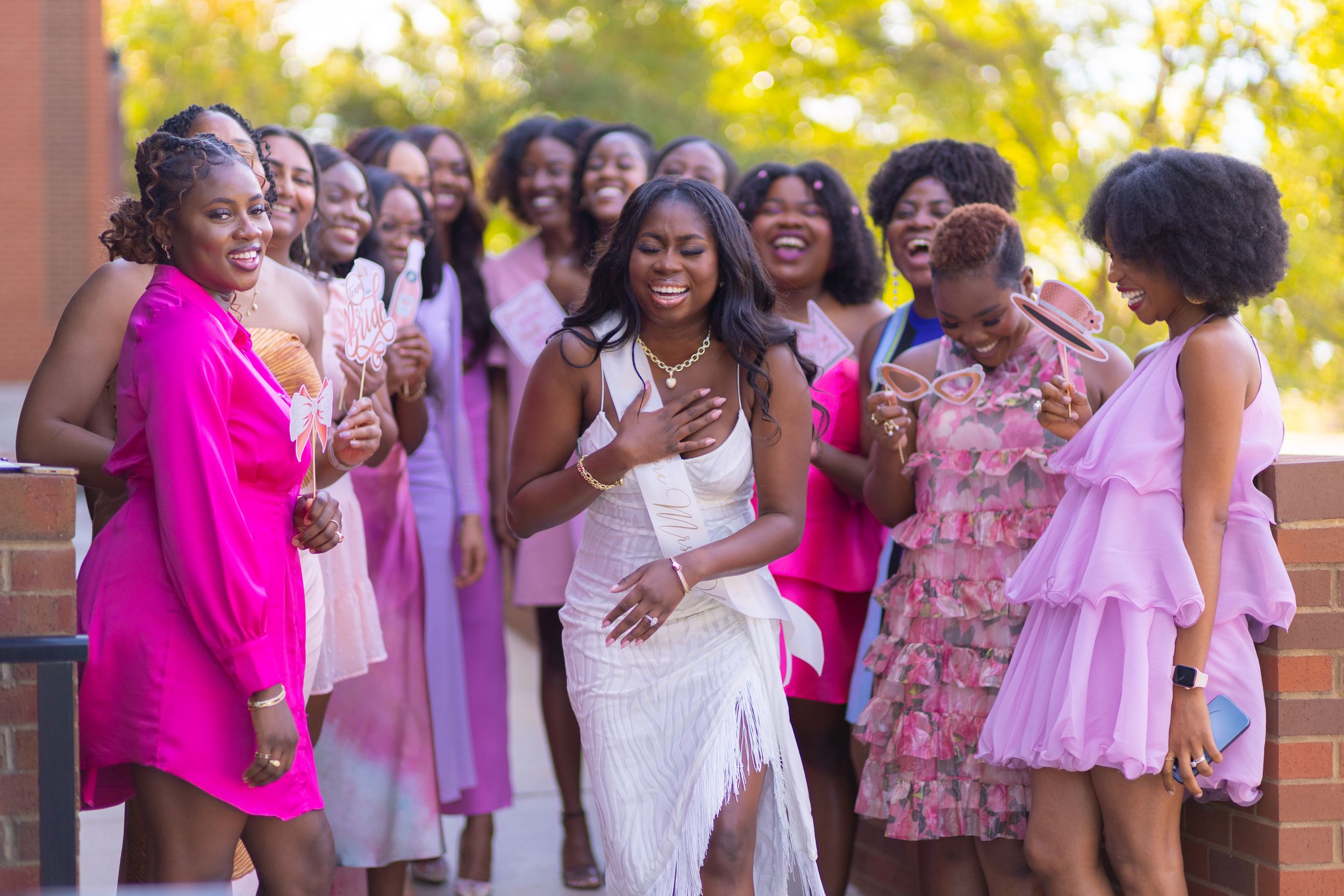 A joyful group of people wearing pink and floral dresses gather outdoors for a celebratory event.