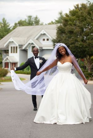 A joyful newlywed couple in formal wedding attire poses together on a driveway with a house in the background.
