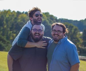 Three people posing together outdoors on a sunny day with trees in the background.