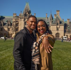 A couple smiles together while posing on the lawn in front of the grand Biltmore Estate on a sunny day.