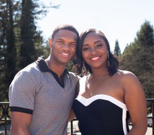 A couple in formal attire smiling outdoors on a sunny day with trees in the background.