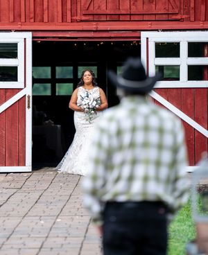 A couple poses for wedding photos in front of a rustic red barn with white trim on a sunny day.