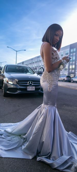 A stunning silver mermaid-style formal gown with lace details sparkles in front of a Mercedes-Benz on a city street.