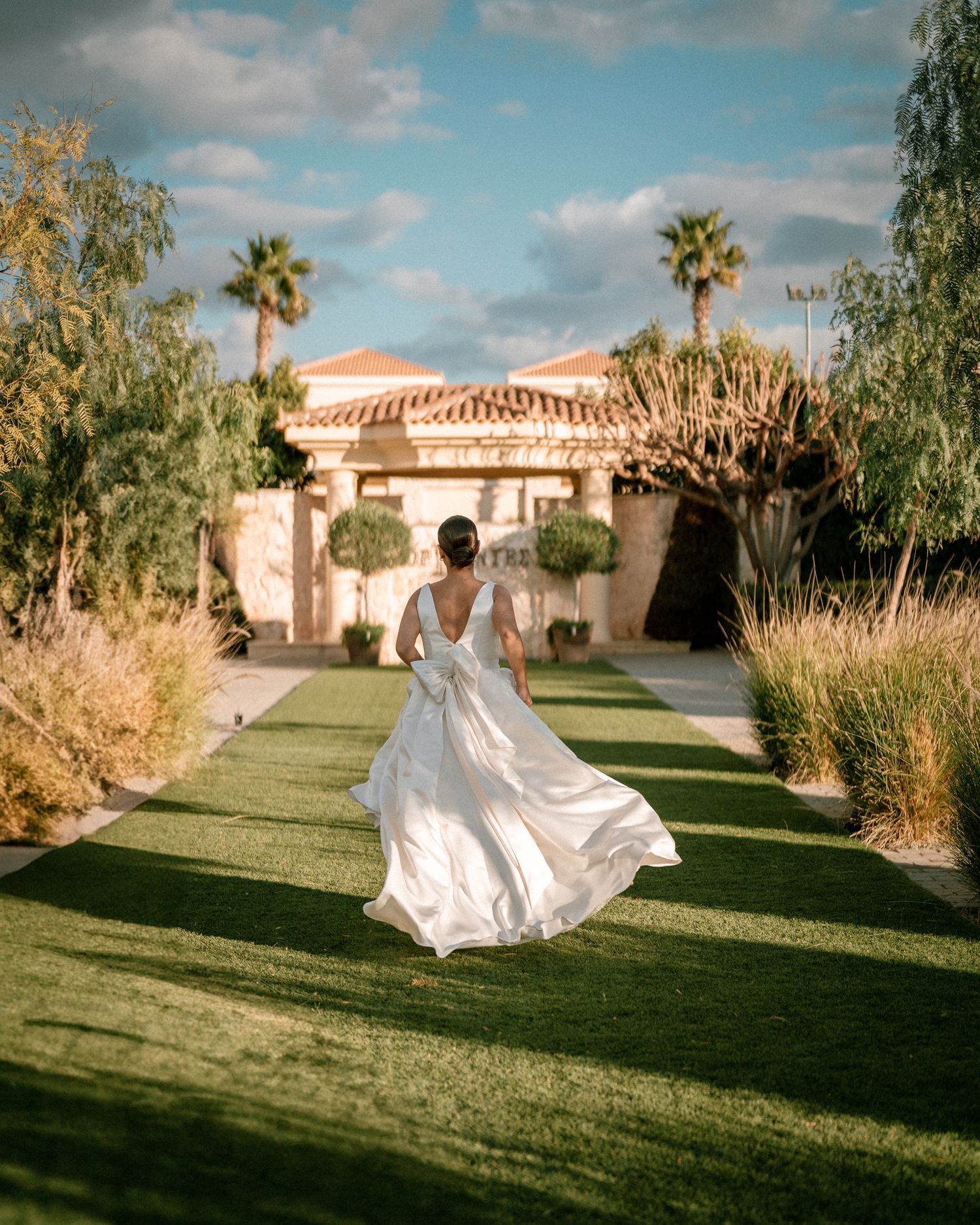 Wedding dress flowing in motion as figure walks toward Spanish-style villa with palm trees.