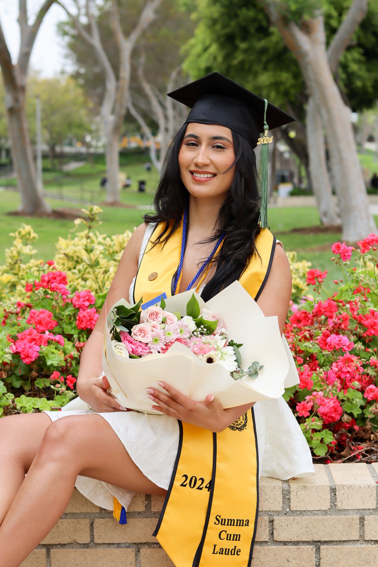 Graduate in cap and gown holding pink flower bouquet in garden with Summa Cum Laude sash.