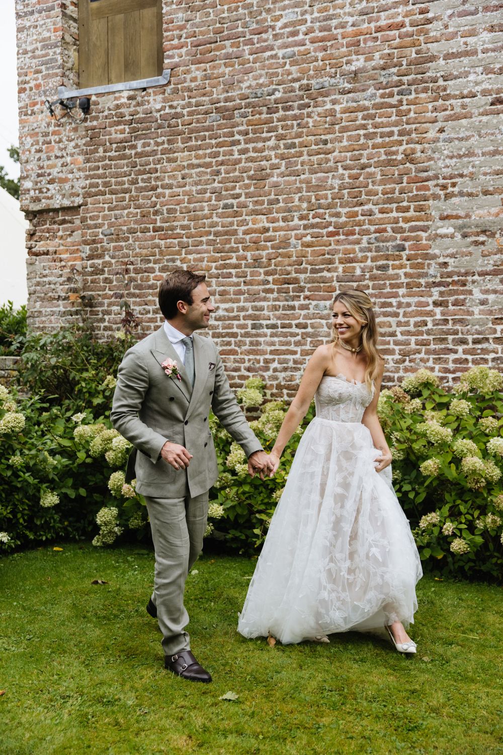 A couple poses playfully by a brick wall with hydrangeas in their wedding attire.