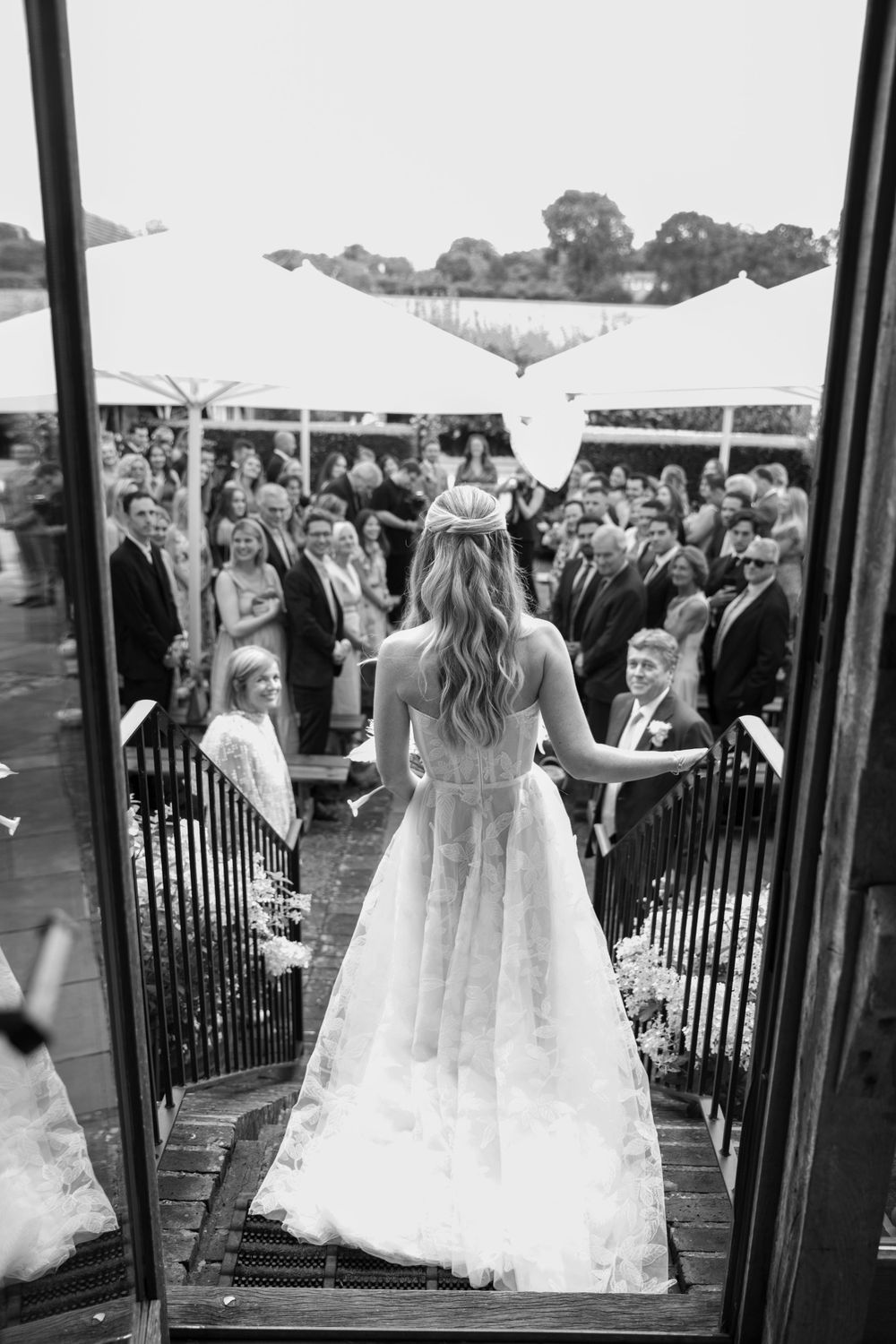 A bride in a flowing wedding gown stands at the top of stairs overlooking wedding guests.