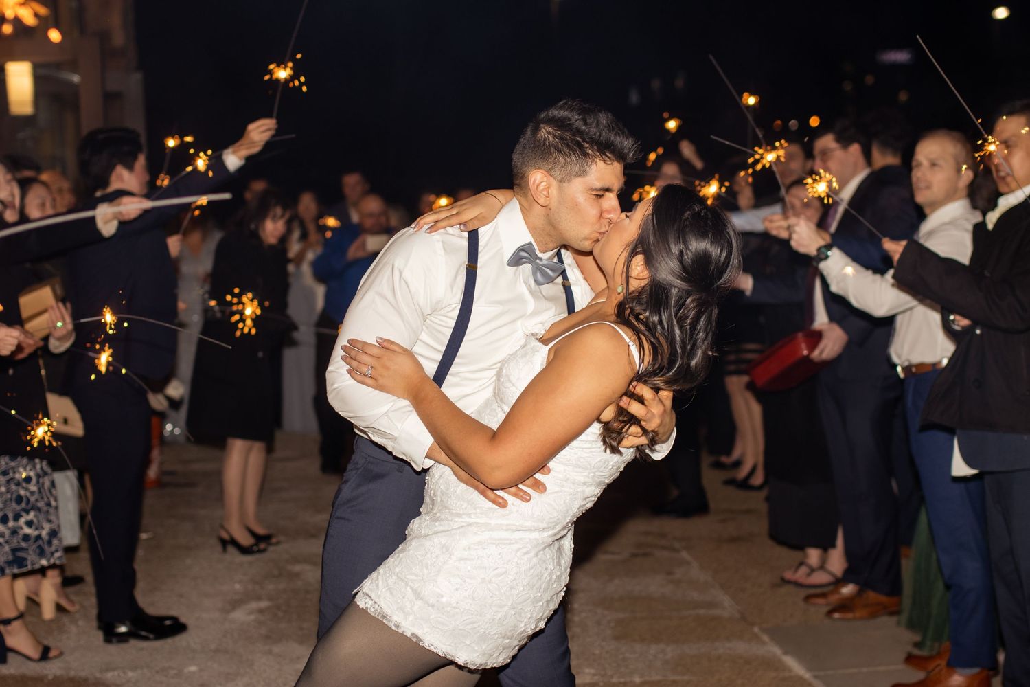 A couple shares a romantic dip kiss during their wedding sparkler sendoff at nighttime.