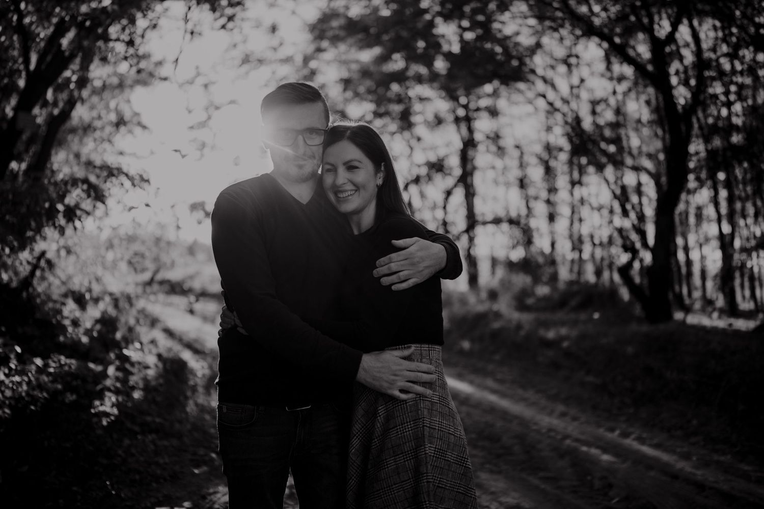 A silhouetted couple embraces on a forest path with sunlight streaming through trees.