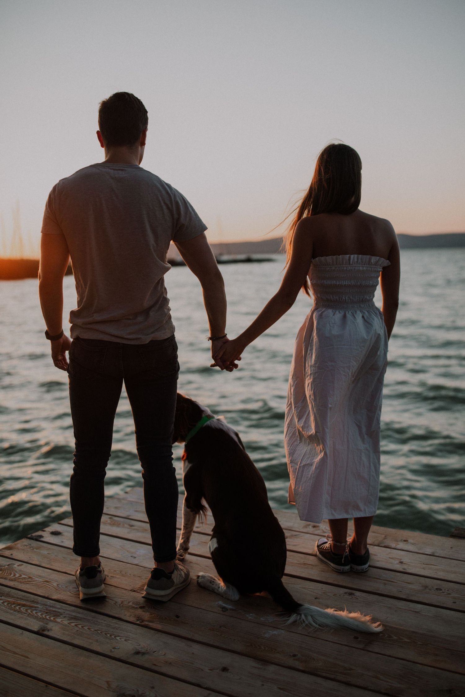 A couple stands on a dock at sunset with their dog gazing at the water.