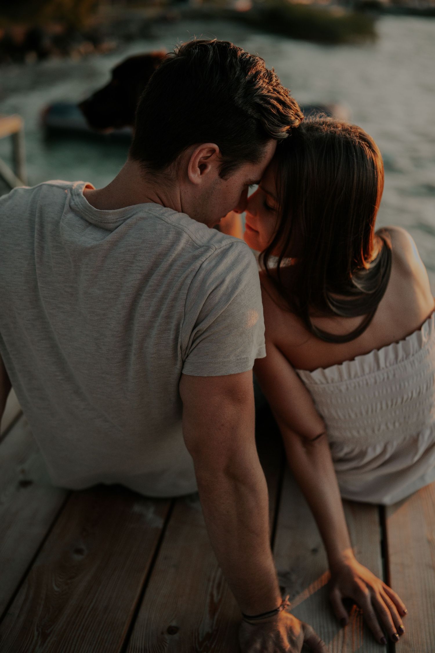 Intimate couple shares a tender moment sitting on wooden dock during golden hour sunset.