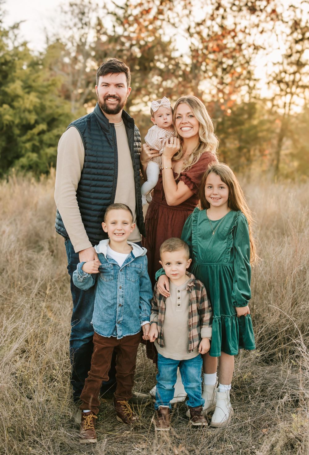 A family of five poses together on an autumn hillside at sunset among tall grasses and evergreen trees.