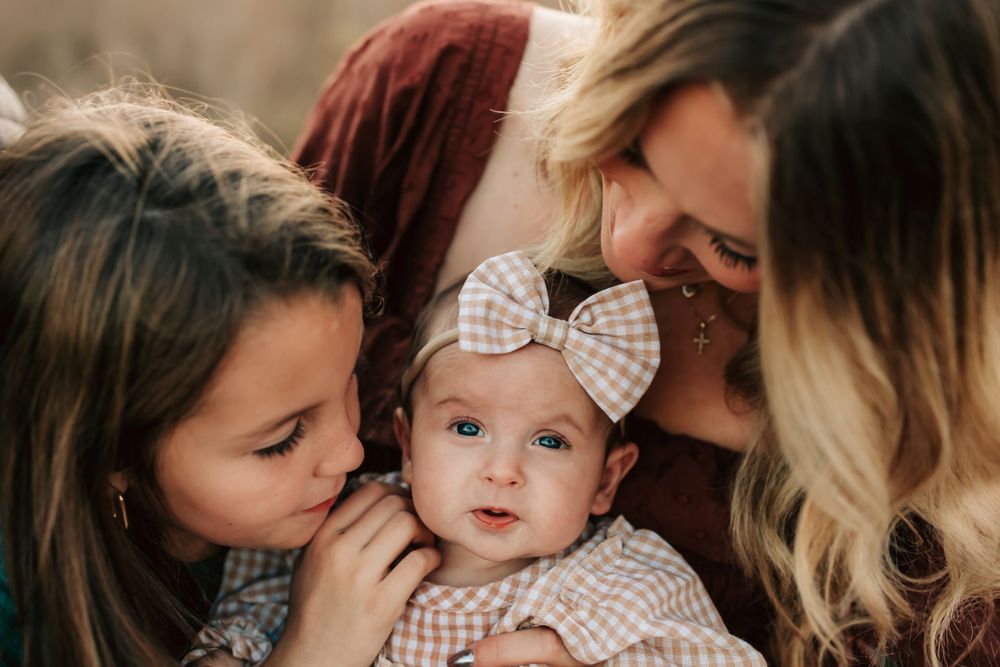 A baby wearing a bow headband is surrounded by loving caretakers in an intimate family portrait.