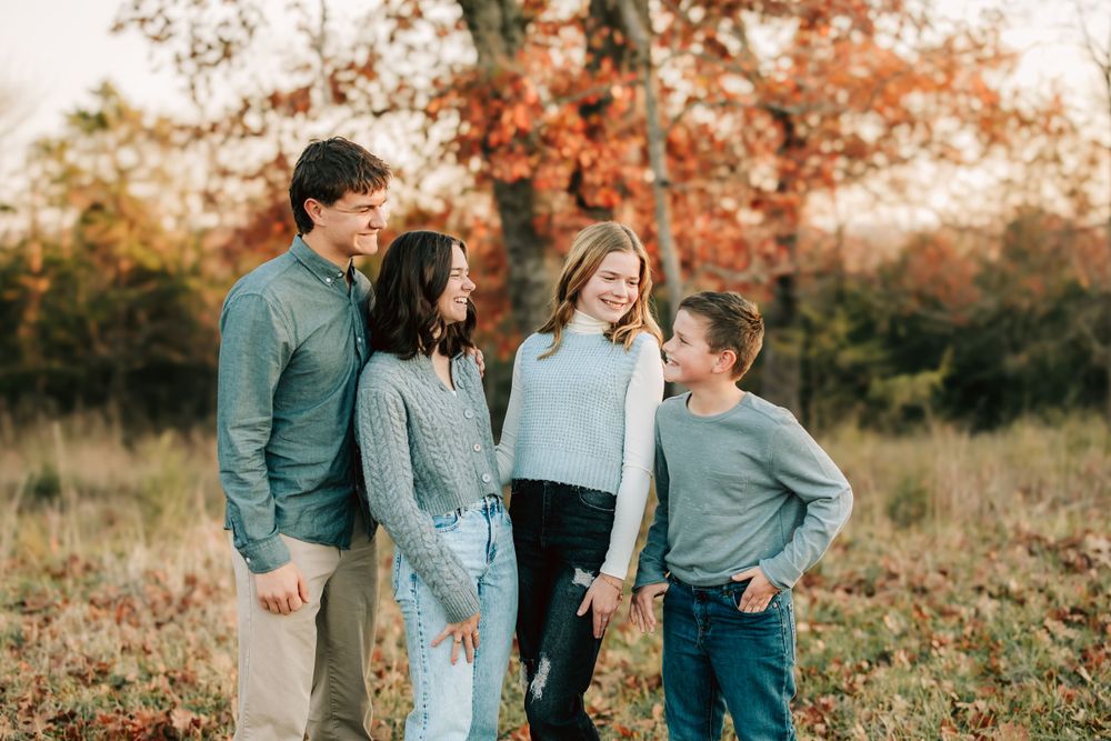 A family of four stands together in a natural outdoor setting with autumn foliage in the background during golden hour.