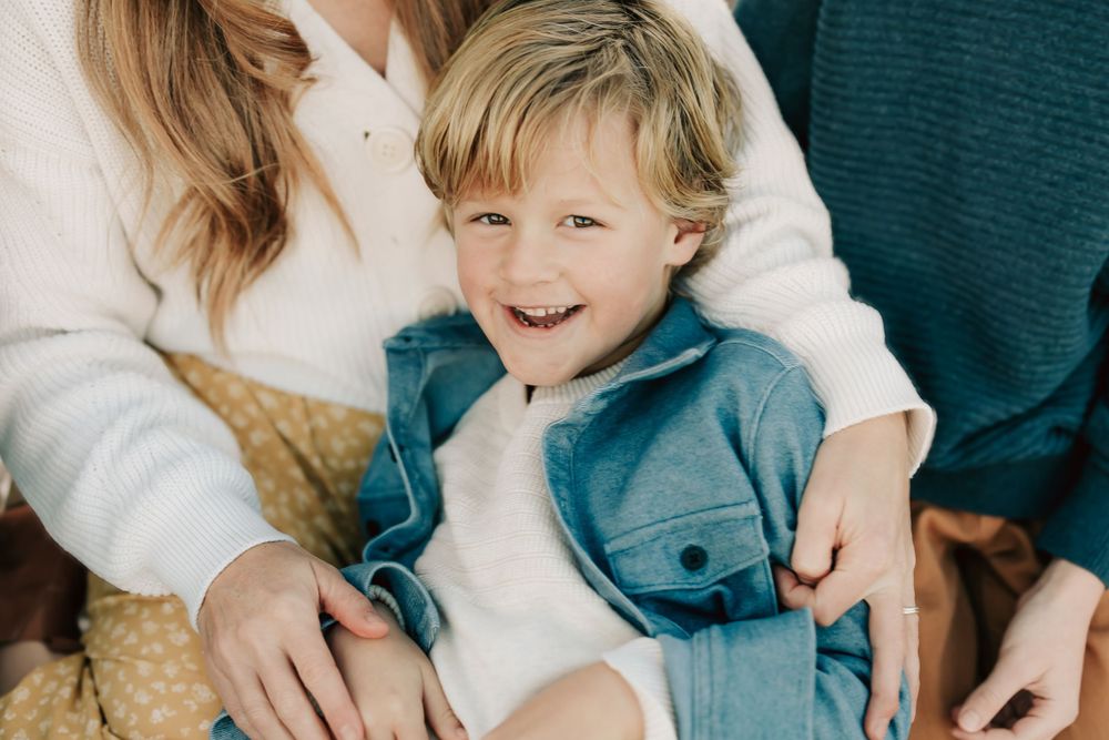 A child wearing a light blue denim jacket smiles brightly while sitting with family members in casual attire.