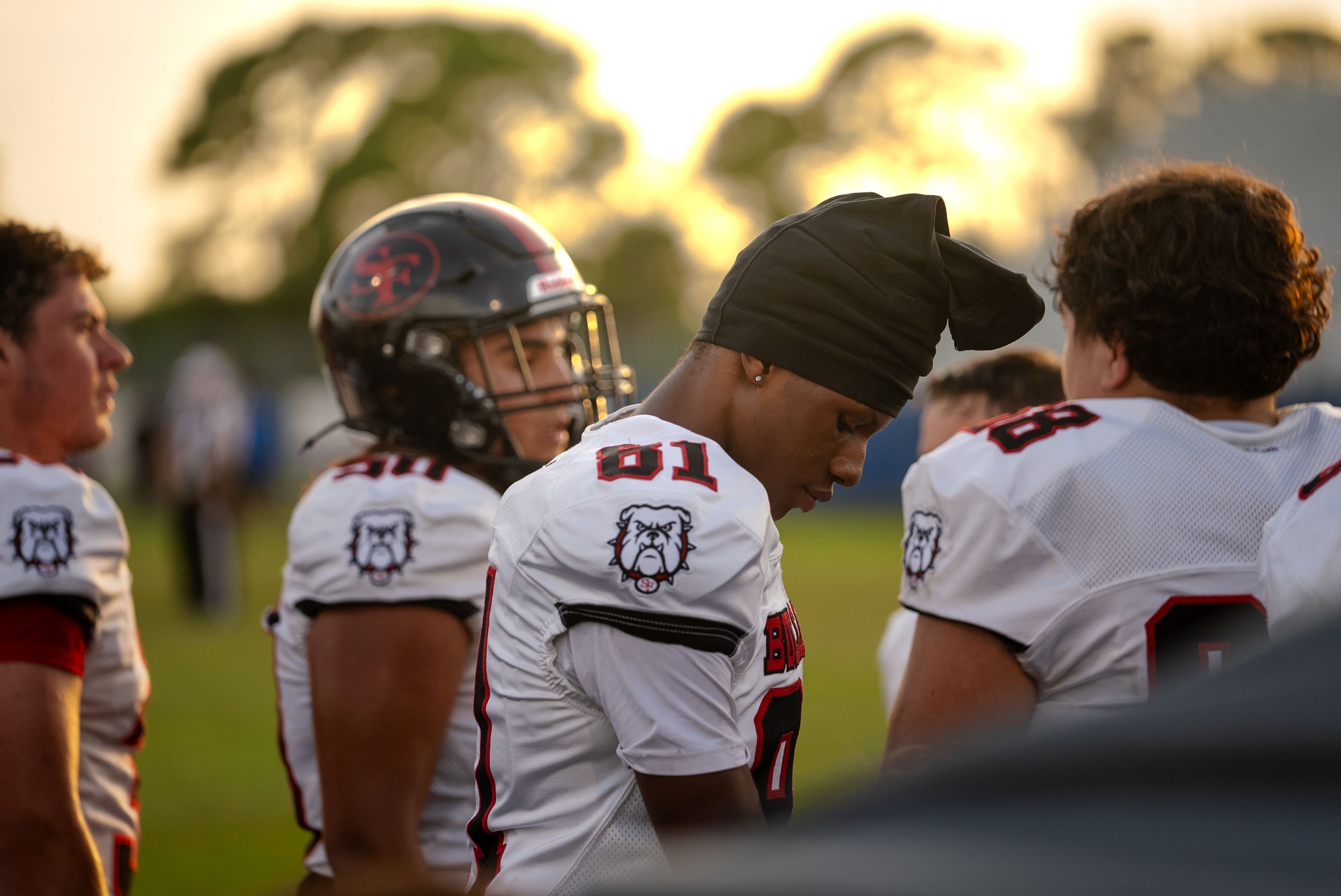 Group of football players in white uniforms gather on sideline at sunset.