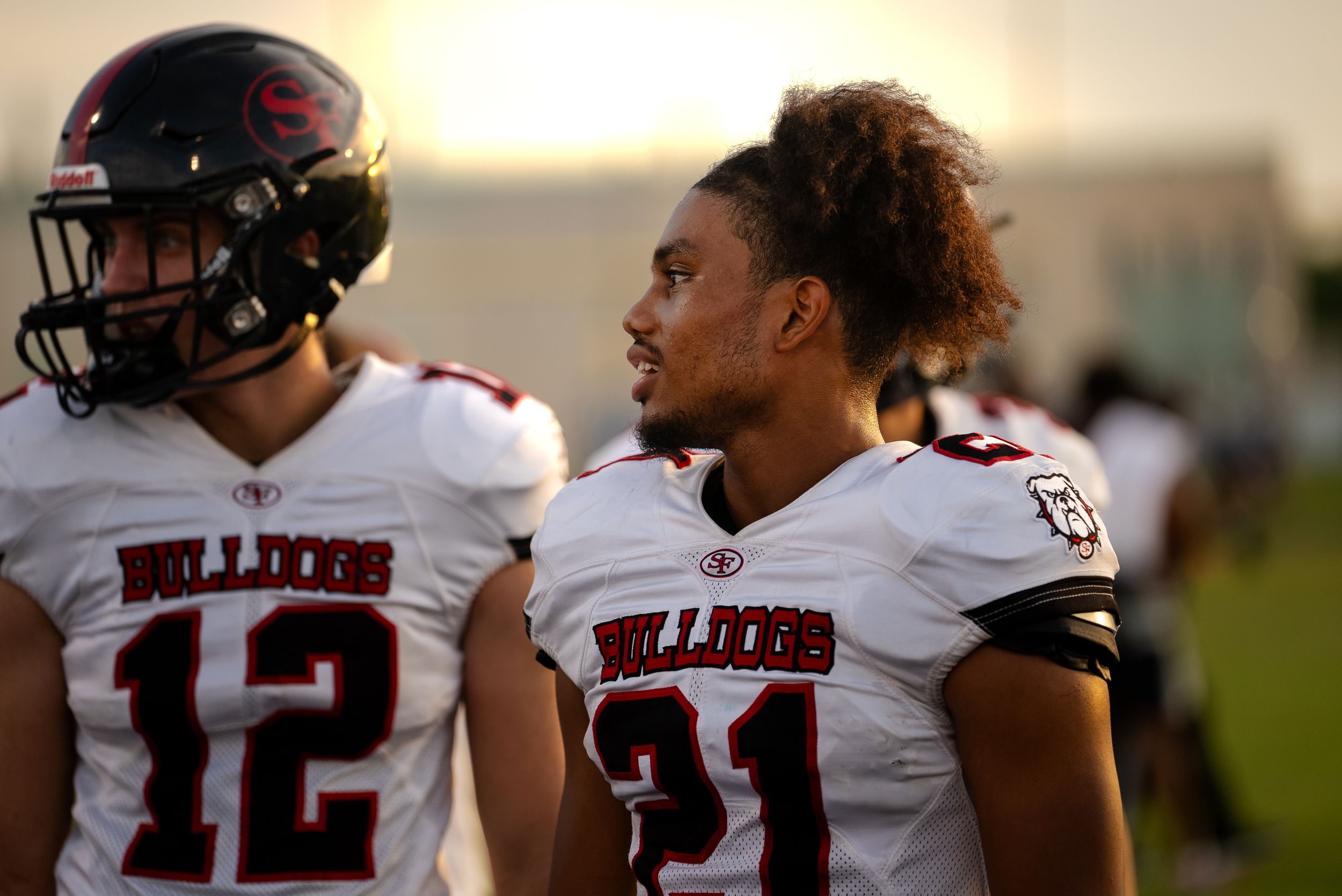 Two football players in white uniforms stand on sideline during sunset.