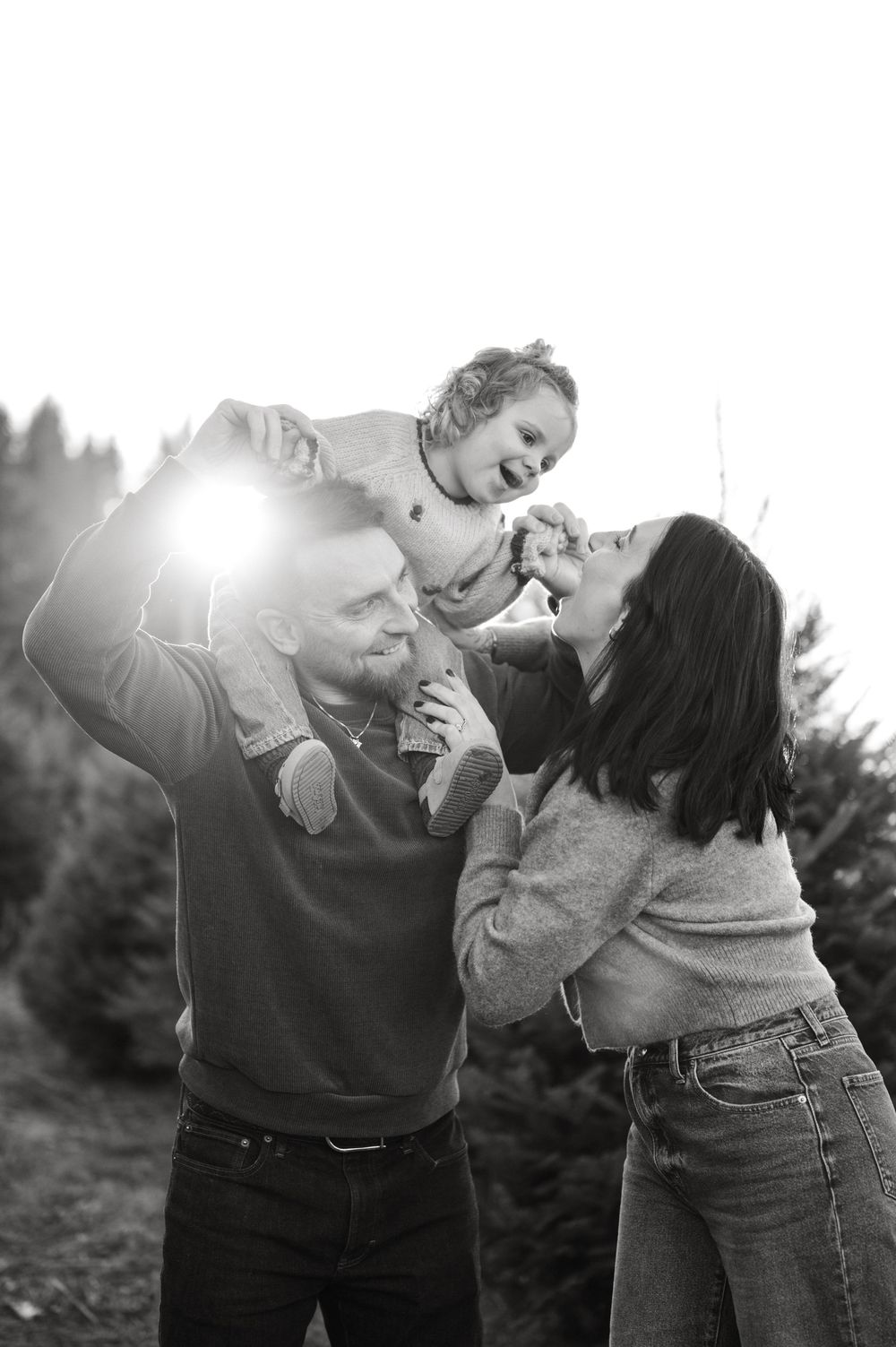 Black and white photo of a joyful family with parents lifting their laughing baby outdoors at a Christmas tree farm.