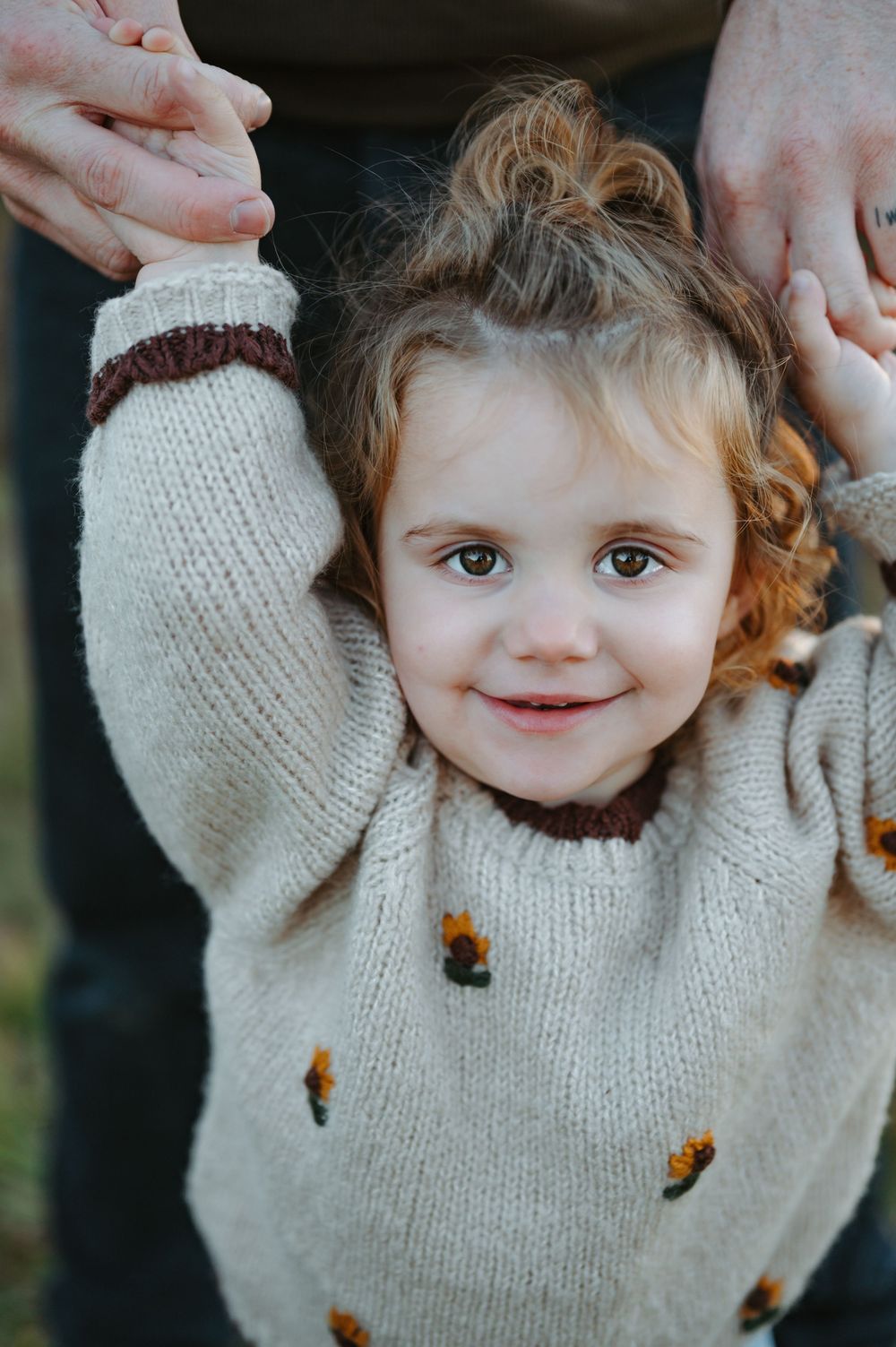 Smiling toddler girl in a cream knit sweater with floral details, arms raised, held by an adult's hands.