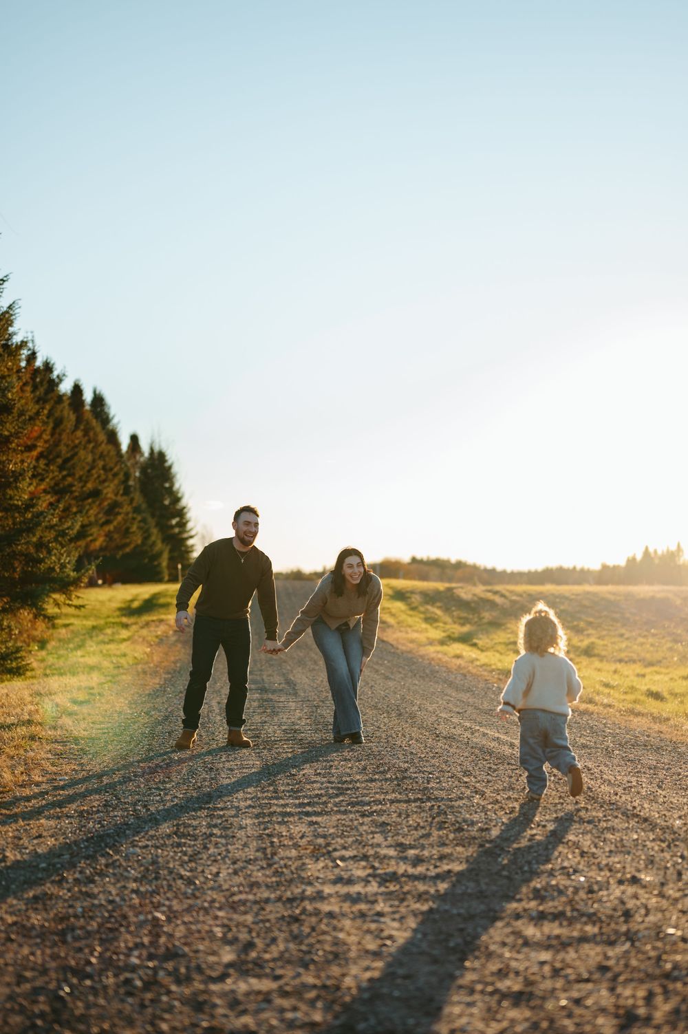 Family of three walking on a sunny dirt road, parents holding hands as toddler runs toward them.