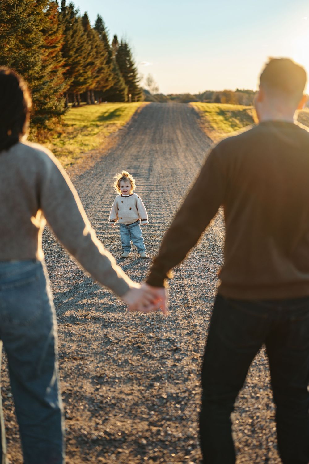 Two parents hold hands on a gravel road at sunset while their toddler walks toward them surrounded by trees.
