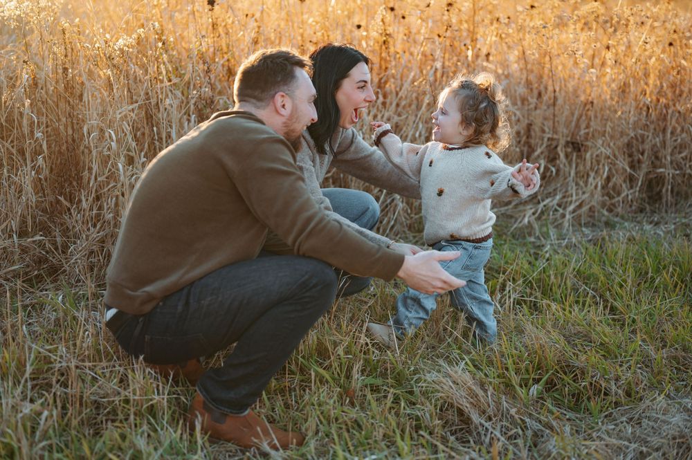 Family with toddler enjoying golden hour in autumn field, parents crouching to interact with young child.