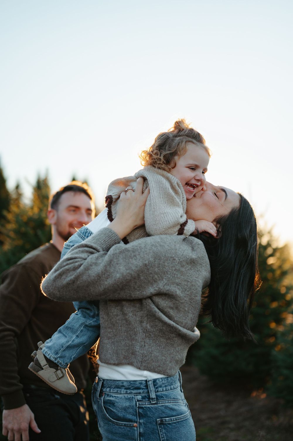 Mother joyfully lifting laughing toddler at a Christmas tree farm.