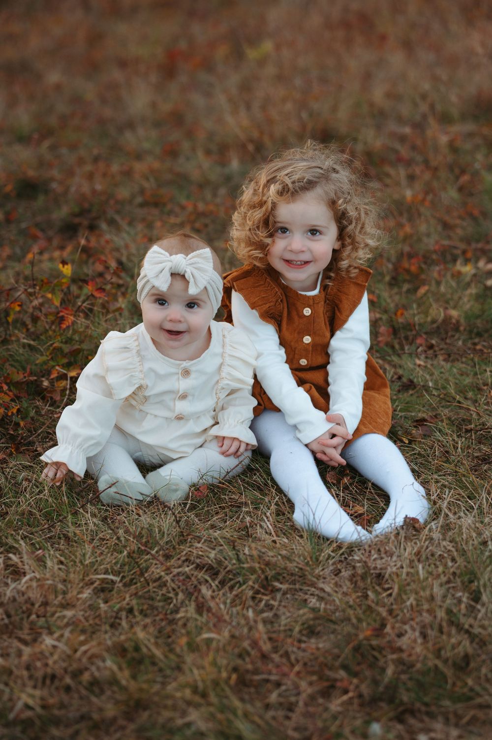 Two children in vintage-inspired clothing sit together on brown grass during an outdoor photo session.