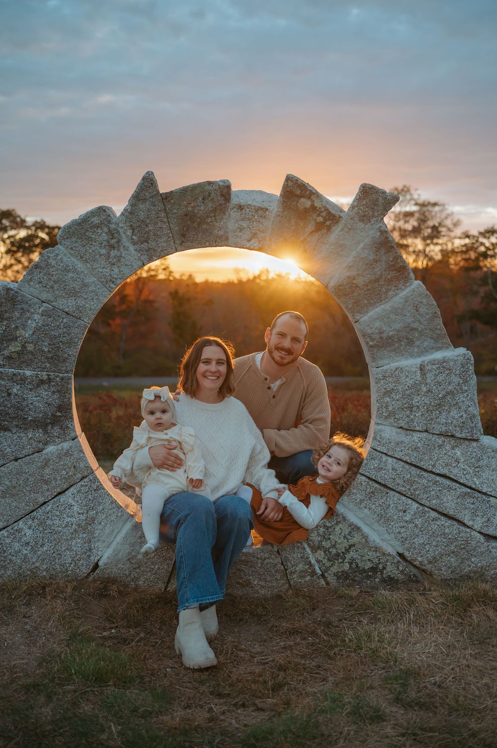Family sits on rocky outcrop during golden hour sunset with mountains in background.