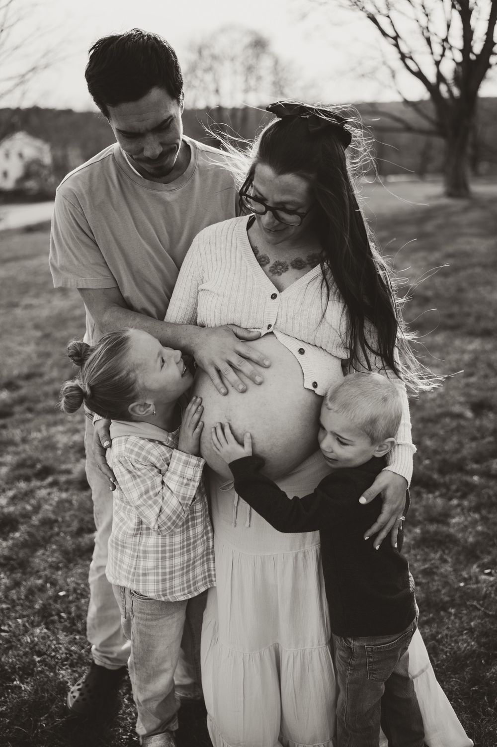 Black and white photo of pregnant woman with husband and two children hugging her baby bump outdoors.