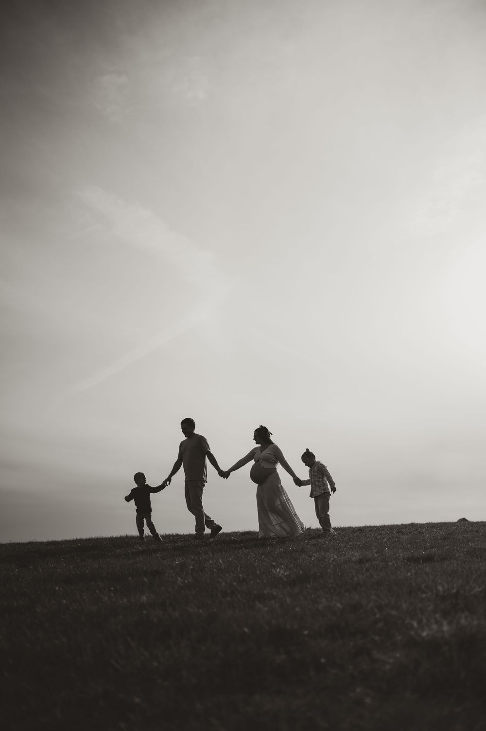 Silhouette of a pregnant family holding hands on a grassy hill at dusk in black and white.