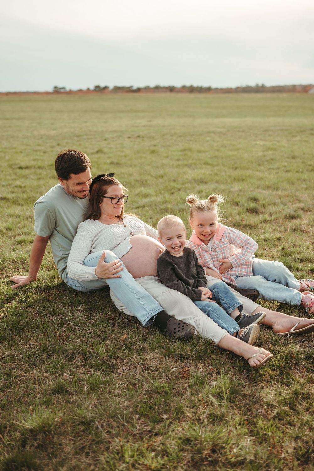 Happy family with pregnant mother and two children sitting on grass field during golden hour.