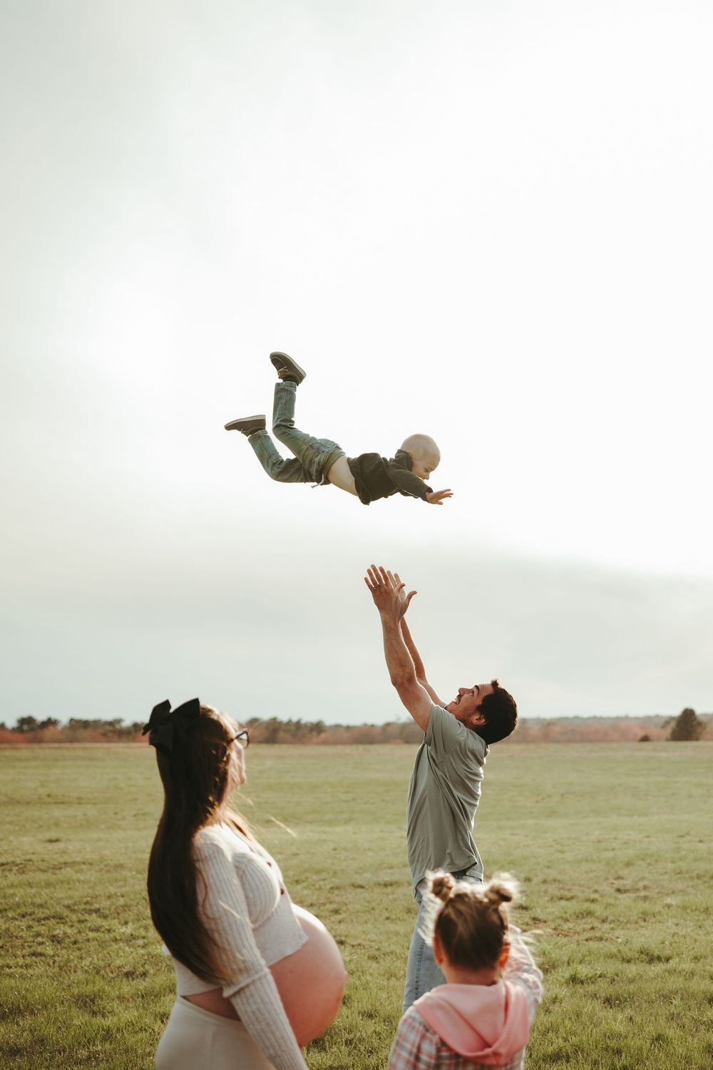 Father tossing child in the air while pregnant mother and daughter watch in an open field.