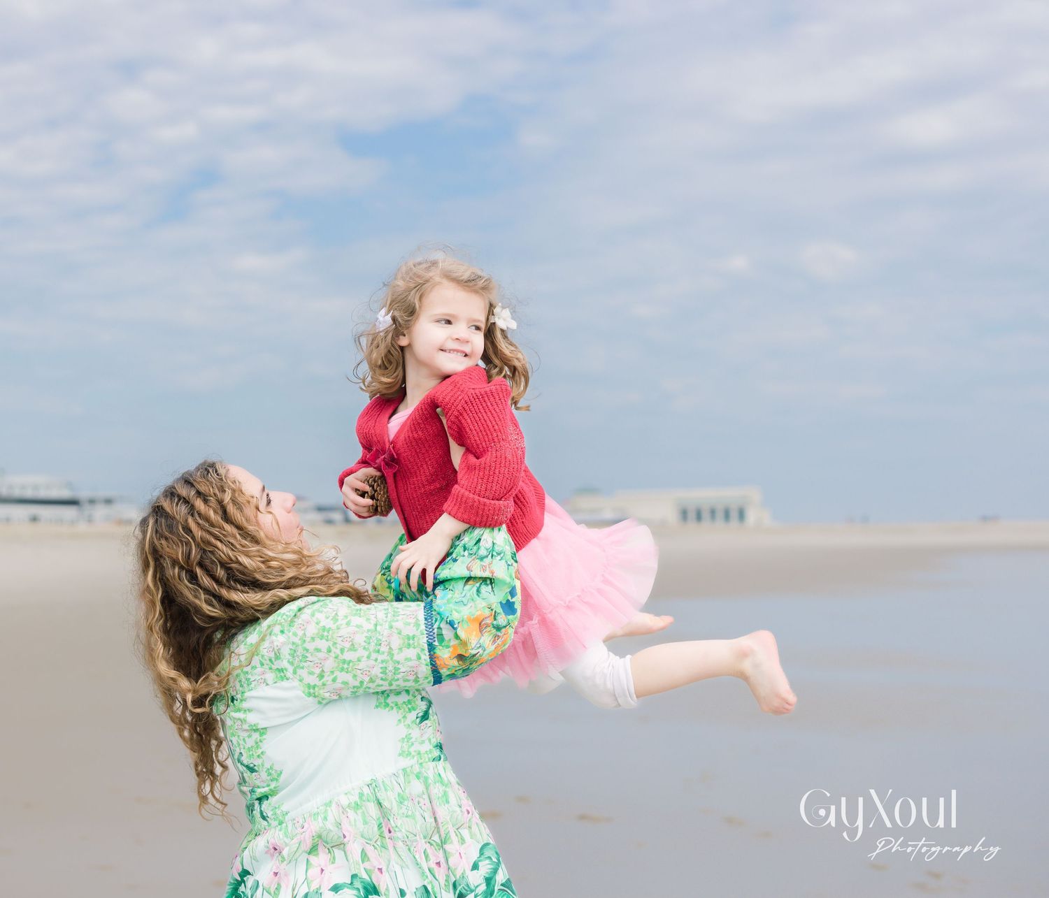 A playful moment captured on a sunny beach as a child in a pink tutu gets lifted into the air against a cloudy sky.