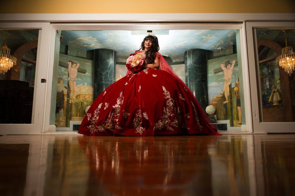 Ornate red ball gown photographed in grand hall with decorative pillars and murals.