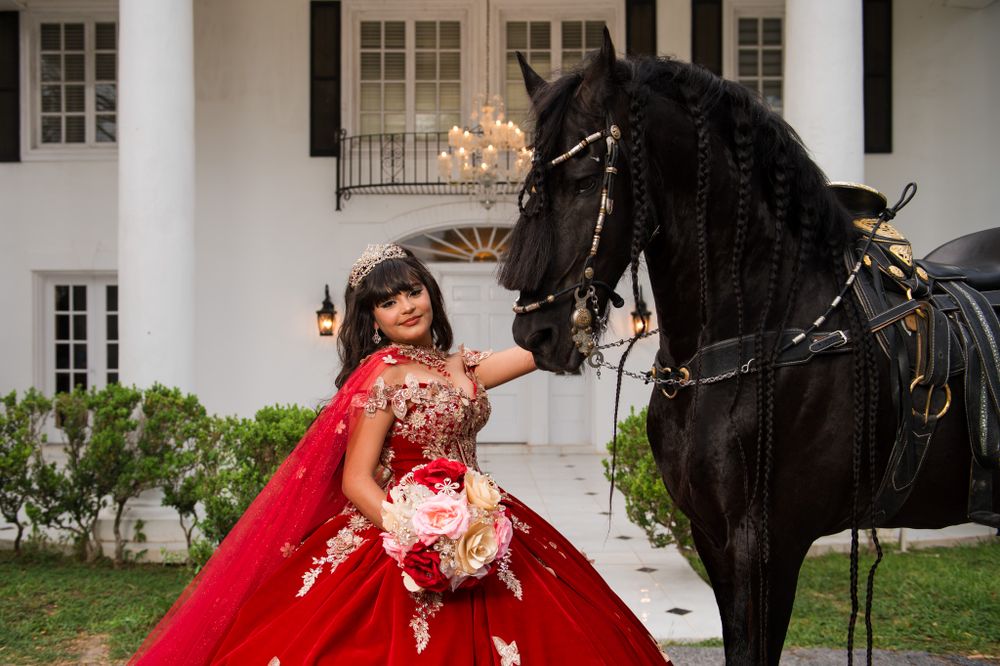 A majestic black horse poses beside someone wearing a red quinceañera dress outside a white mansion.