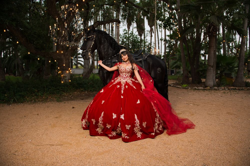 Red quinceañera dress with gold embroidery photographed in outdoor garden setting.