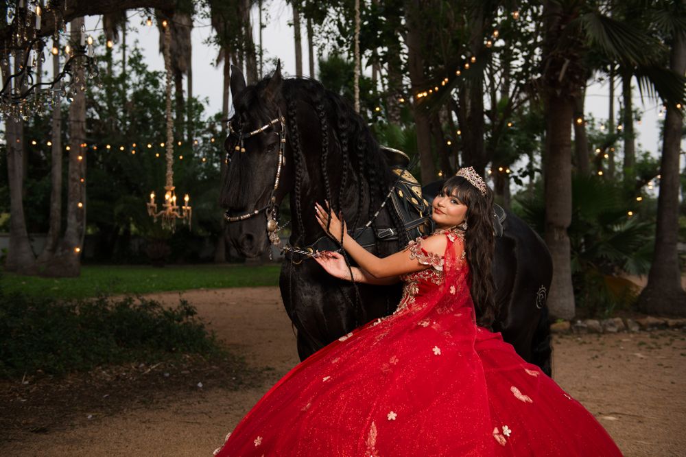A stunning red quinceañera dress glimmers in an enchanted forest setting with string lights and a black horse.