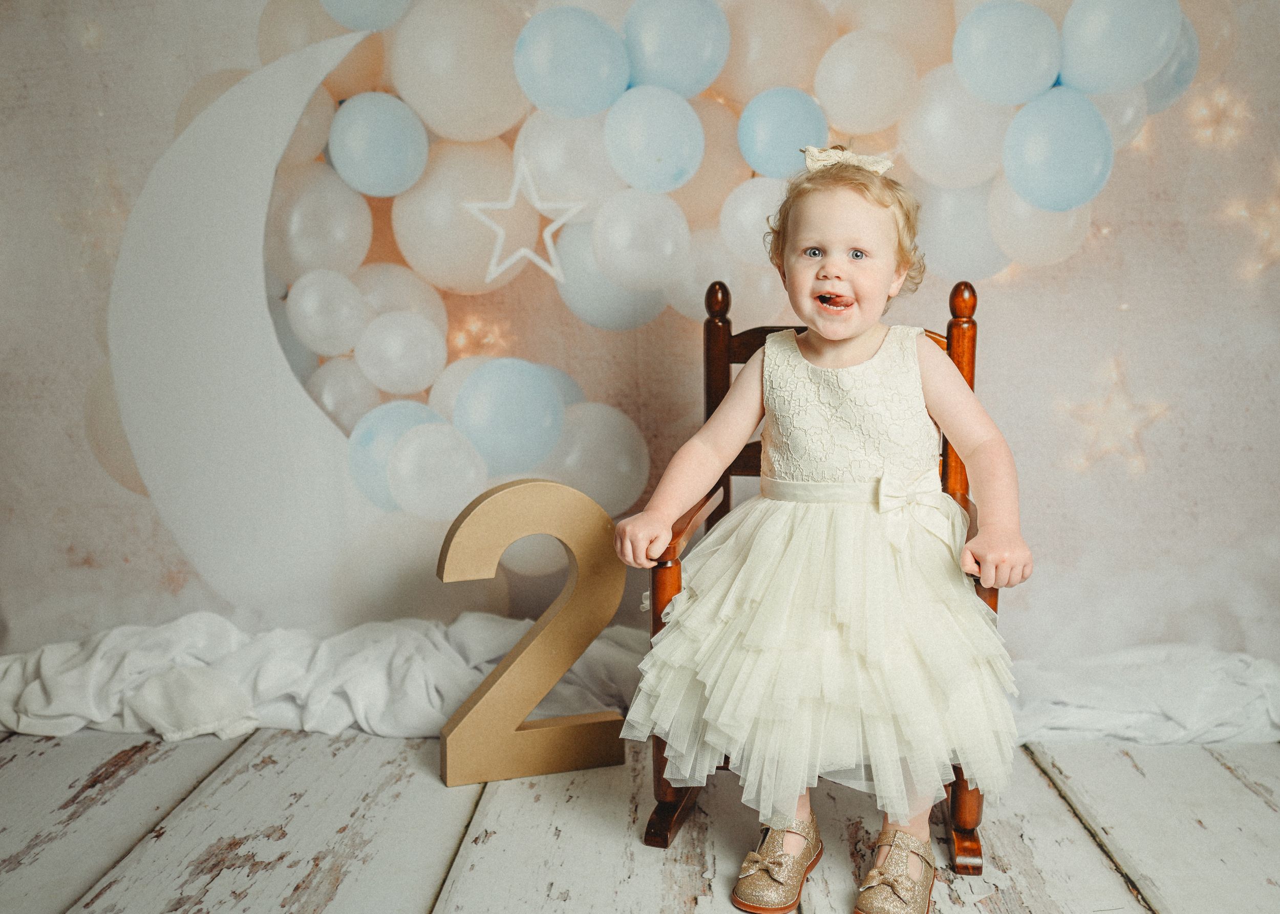 Toddler in white tulle dress sitting on wooden chair with birthday decorations.