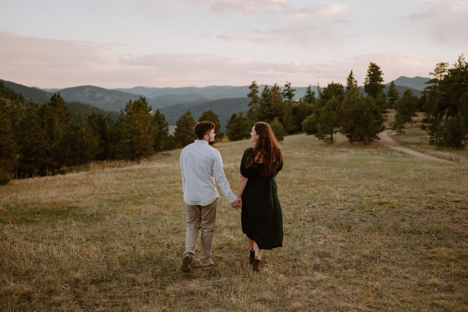 A couple holding hands walks through a grassy field towards a forested mountain vista under a cloudy sunset sky.