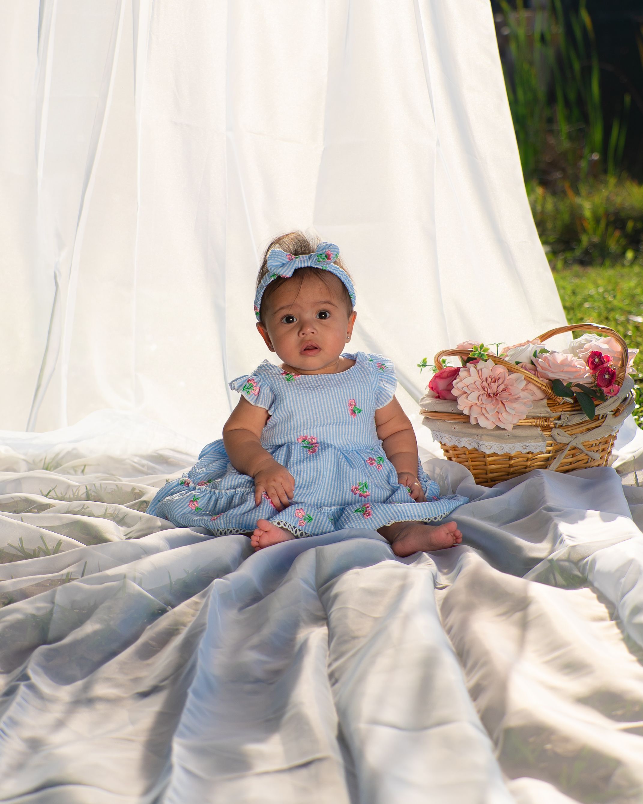 Adorable baby girl in a blue floral dress sitting on a blanket with a flower basket.