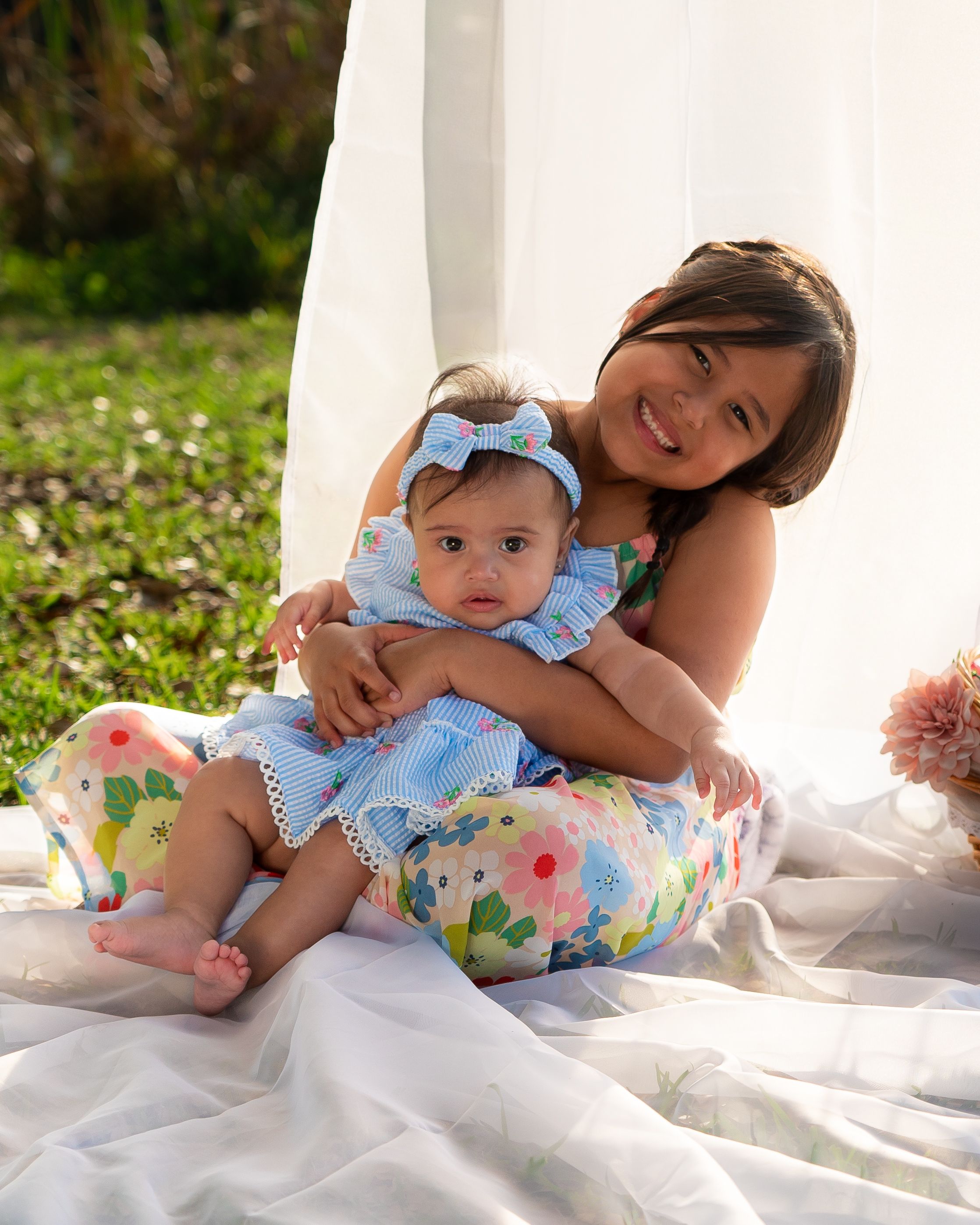 Older sister hugging baby sibling outdoors on a blanket in matching floral outfits.