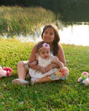 Two sisters sitting together on green grass near a pond surrounded by pink flowers.