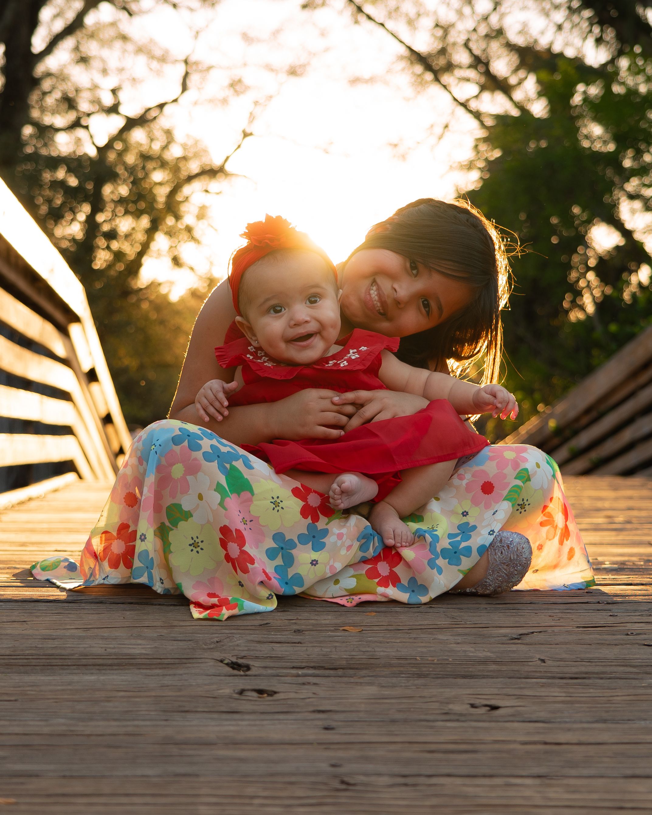 Older sister hugging baby in red outfit on a wooden bridge at sunset.