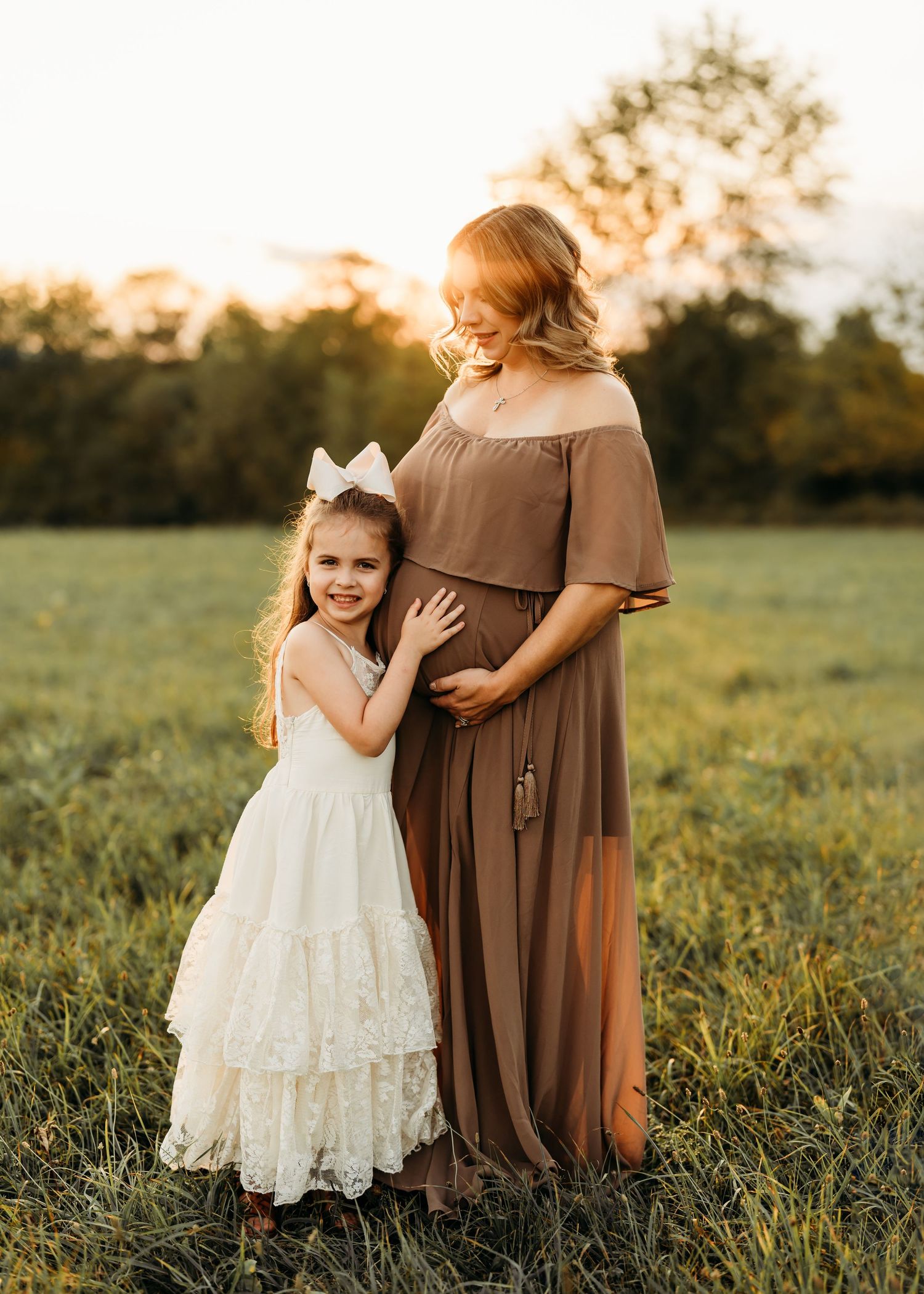 Heartwarming maternity moment with young girl in white dress embracing bump in sunlit field.