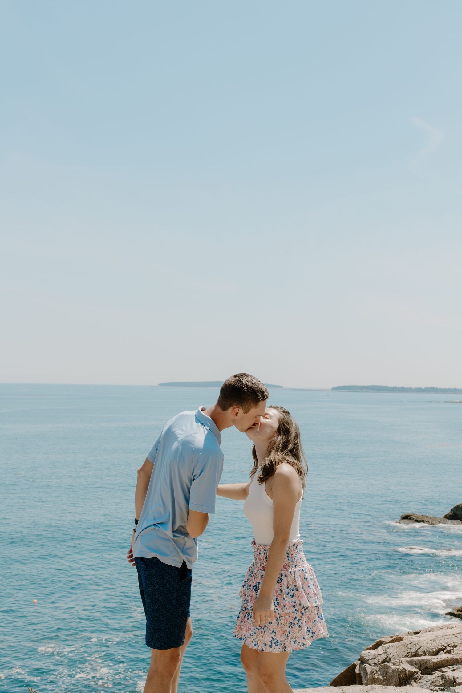 Young couple sharing a kiss on rocky coastal overlook with vivid blue ocean in the background.