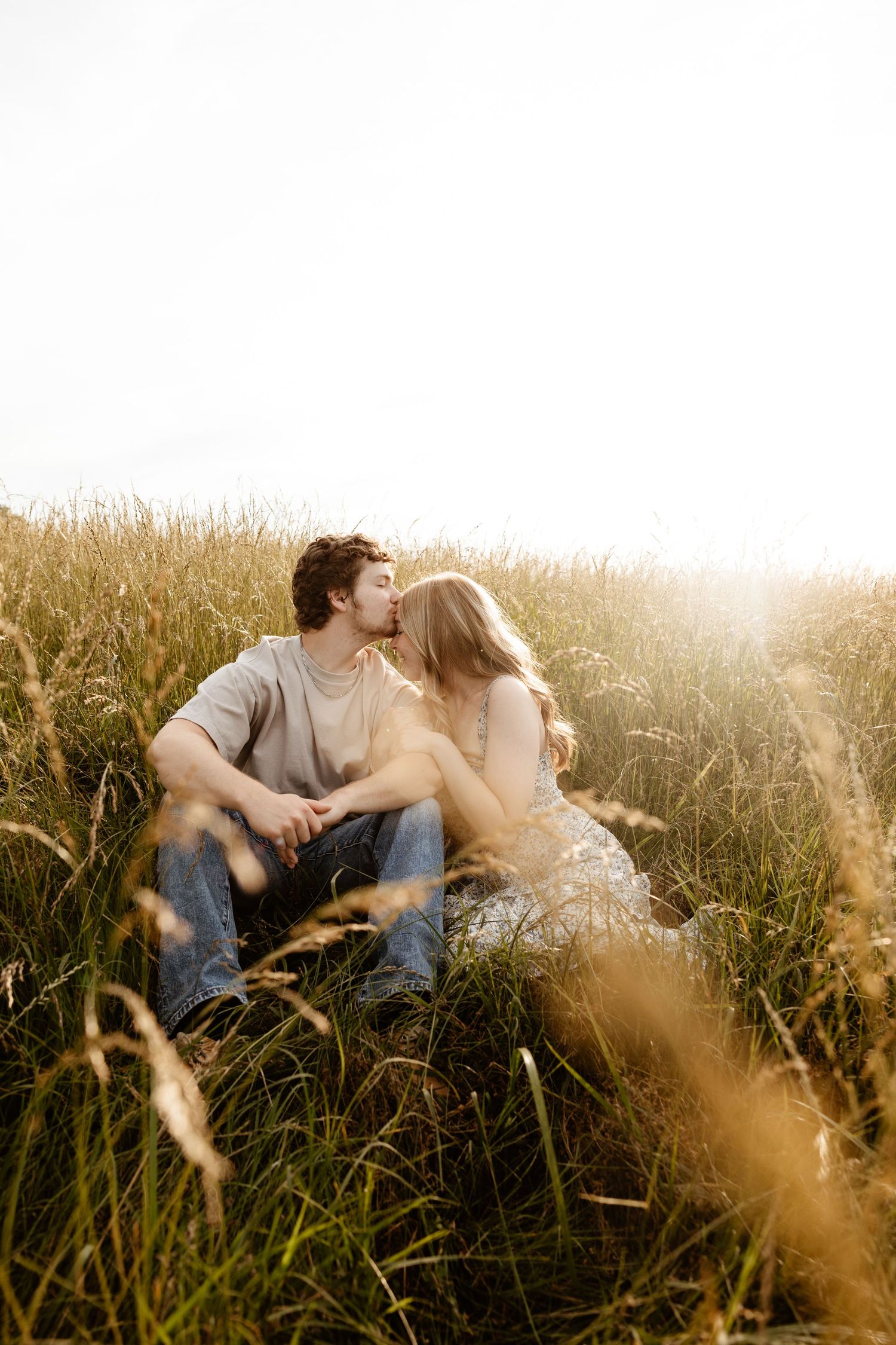 Couple sharing a kiss while sitting together in a sun-drenched golden grass field at sunset.